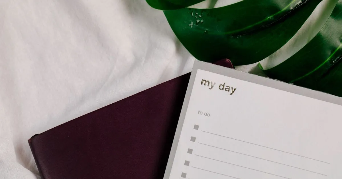 A close-up of a to-do list notebook with lush green leaves on a white textile surface.